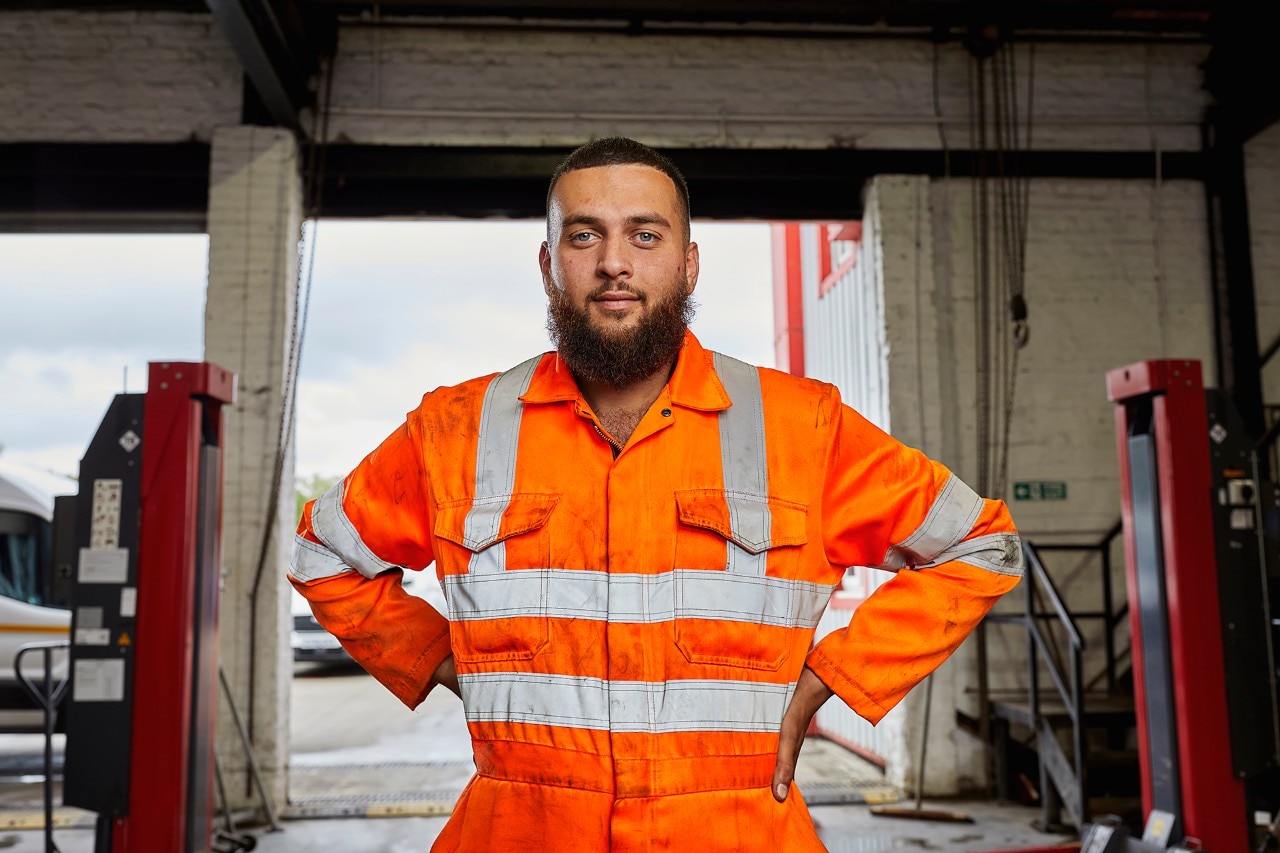Enterprise Mobility Automotive Technician Mechanic Apprentice in the UK dressed in high visibility overalls smiling and with hands on hips. 