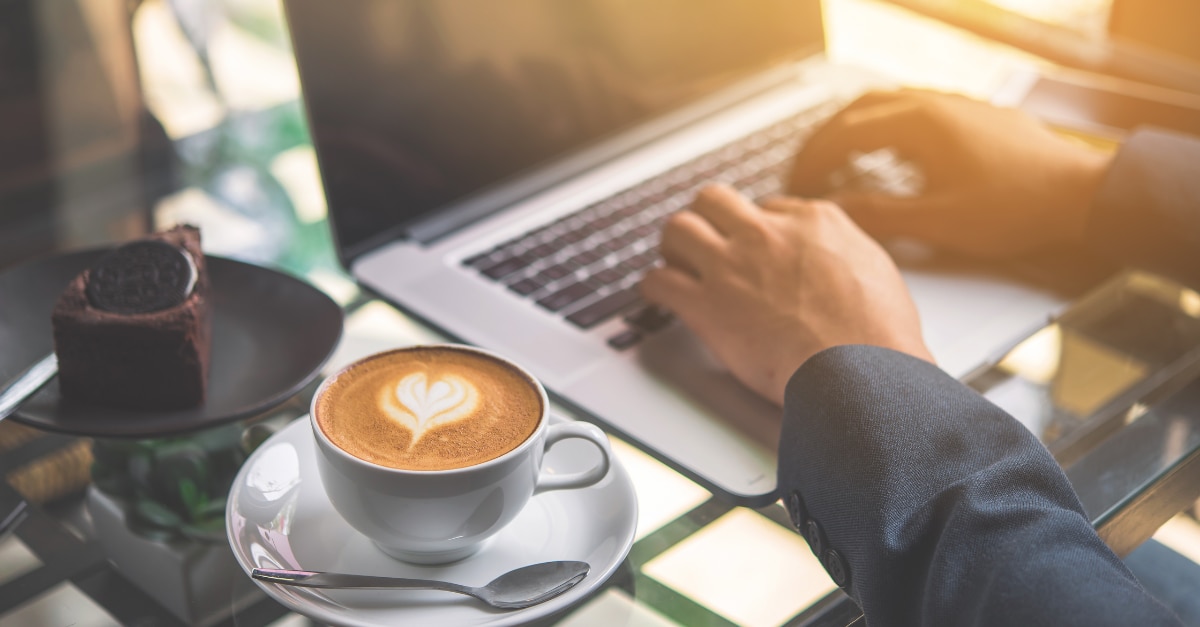 Hands typing on a Macbook with coffee and cake on table