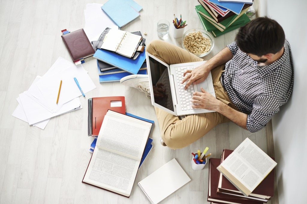 Student sitting on the floor with a laptop in his lap surrounded by notebooks, books, and papers