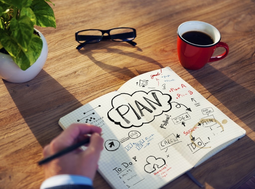 Hand writing in notebook with plans on a desk with a plant, glasses, and cup of coffee
