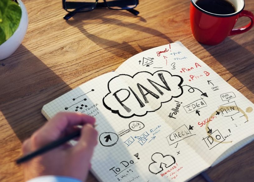 Hand writing in notebook with plans on a desk with a plant, glasses, and cup of coffee