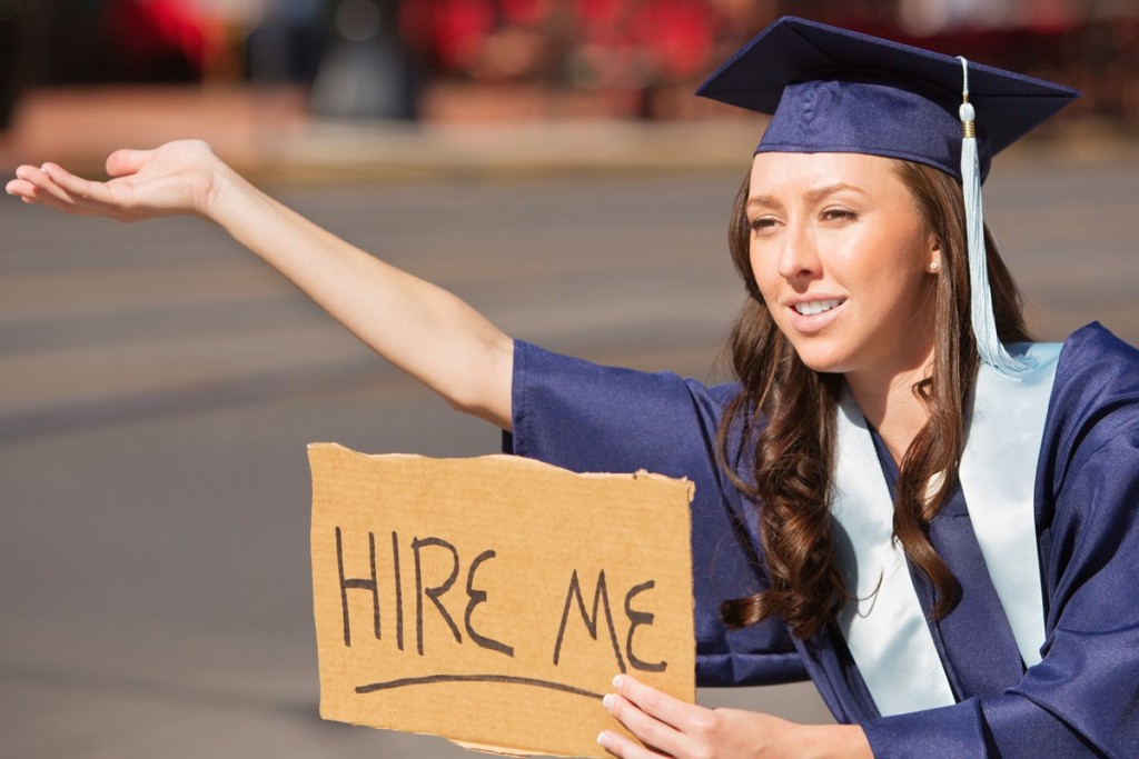 Student in graduation cap and gown holding cardboard sign that says "hire me"
