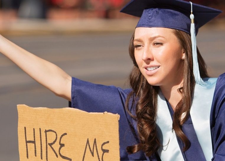 Student in graduation cap and gown holding cardboard sign that says "hire me"
